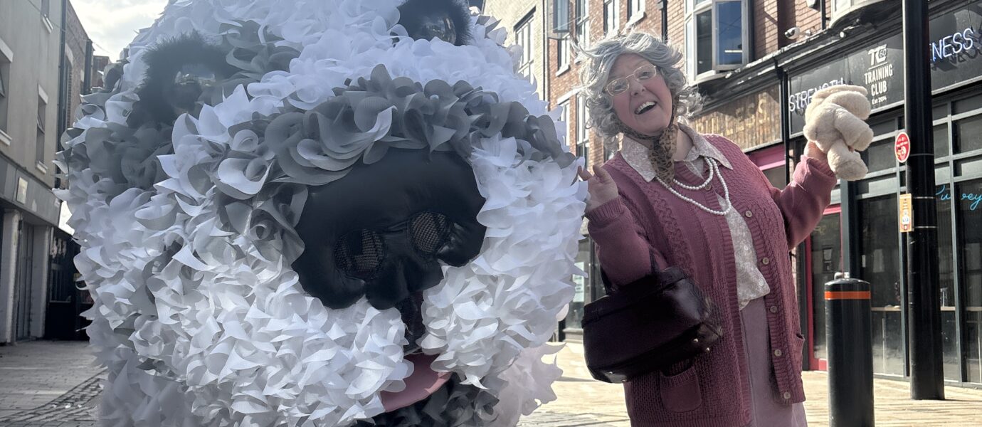 A woman dressed in pink joyfully poses next to a large, fluffy dog sculpture made of white and grey materials on a city street with brick buildings in the background.