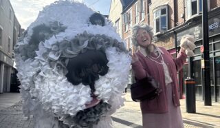 A woman dressed in pink joyfully poses next to a large, fluffy dog sculpture made of white and grey materials on a city street with brick buildings in the background.