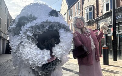 A woman dressed in pink joyfully poses next to a large, fluffy dog sculpture made of white and grey materials on a city street with brick buildings in the background.
