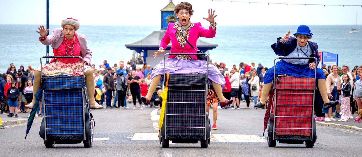 Three people dressed as elderly women in colourful clothes race on large checked shopping trolleys along a seaside street, with a crowd watching and the sea in the background.