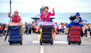 Three people dressed as elderly women in colourful clothes race on large checked shopping trolleys along a seaside street, with a crowd watching and the sea in the background.