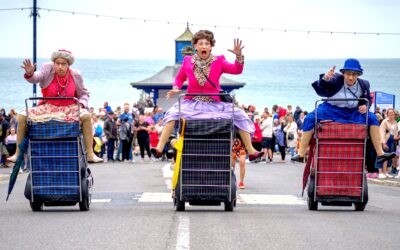 Three people dressed as elderly women in colourful clothes race on large checked shopping trolleys along a seaside street, with a crowd watching and the sea in the background.