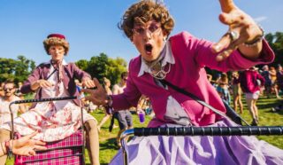 Two people dressed as elderly women in colourful costumes enthusiastically pose outdoors at a lively, sunny event, with one reaching towards the camera and the other pushing a zimmer frame behind. Crowds and green trees appear in the background.