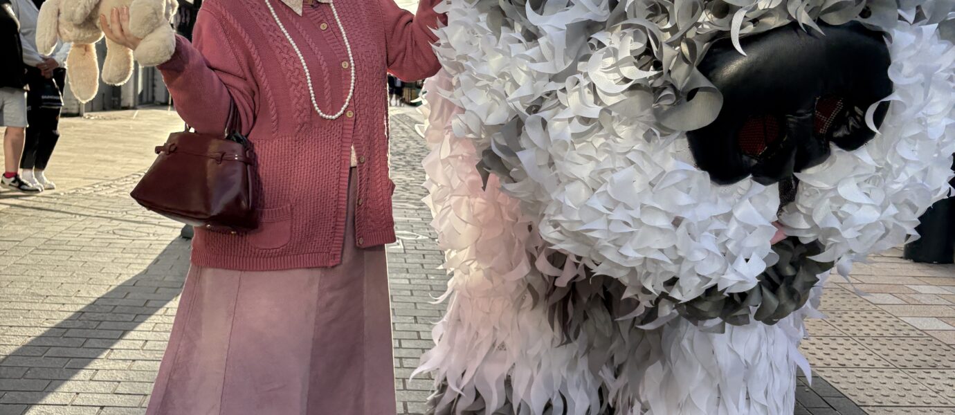 An older woman in a pink outfit poses smiling next to a large, white furry animal puppet on a city street, under a cloudy sky, with buildings in the background.