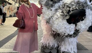 An older woman in a pink outfit poses smiling next to a large, white furry animal puppet on a city street, under a cloudy sky, with buildings in the background.