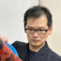 A person with short black hair and glasses, wearing a black jacket, looks intently at a blue object. The background is plain and light-coloured.