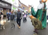 People walk along a busy street with shops. A person in a green wizard costume and another in fantasy attire interact near a man with a dog. Several people watch, some smiling, and a woman in a wheelchair is in the background.