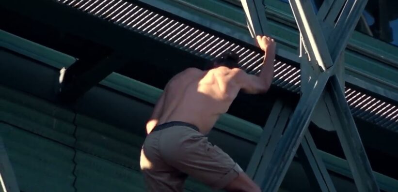 A topless person wearing shorts climbs up the side of a building, gripping a metal structure with one hand and foot visible, against a backdrop of glass windows and beams.