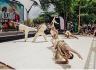 A group of dancers in beige outfits perform expressive movements on an outdoor stage, with a crowd watching and a large banner in the background.