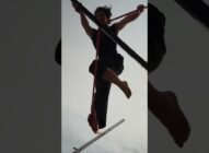 A person balances barefoot on a slackline high above the ground, holding onto the line with both hands and feet, silhouetted against a cloudy sky.