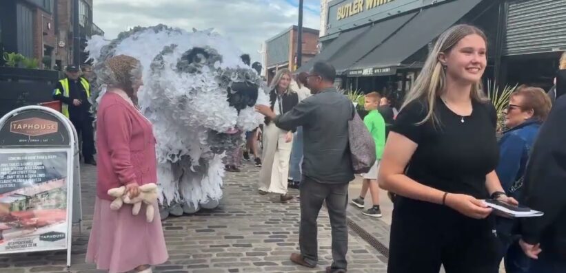A street scene shows people interacting with a large, white polar bear puppet. Some passers-by are smiling whilst others take photos or watch the performance. Shops and a café are visible in the background on a cobbled street.