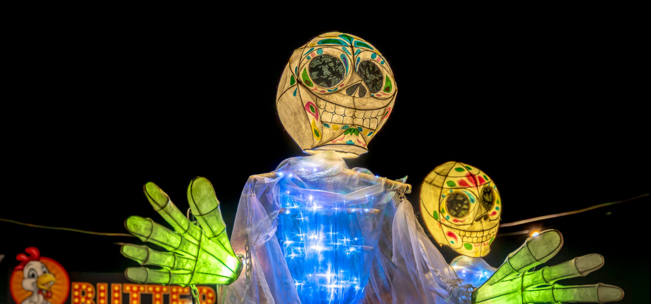 Two colourful, illuminated skeleton figures with big smiles and glowing green hands stand against a dark night sky, with food stall signs visible in the background.