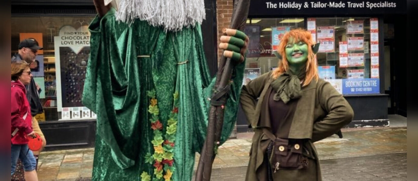 A tall person dressed as a wizard with a green robe and staff stands beside a shorter person in a green fantasy costume, both posing outdoors on a city street.