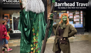 A tall person dressed as a wizard with a green robe and staff stands beside a shorter person in a green fantasy costume, both posing outdoors on a city street.