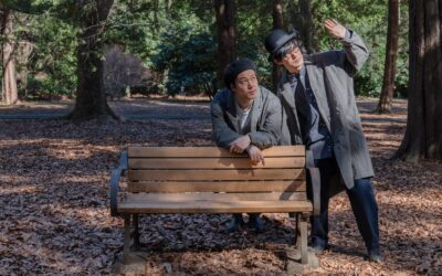 Two men in hats and coats stand by a wooden park bench, with one man leaning on the backrest and the other gesturing towards the sky.
