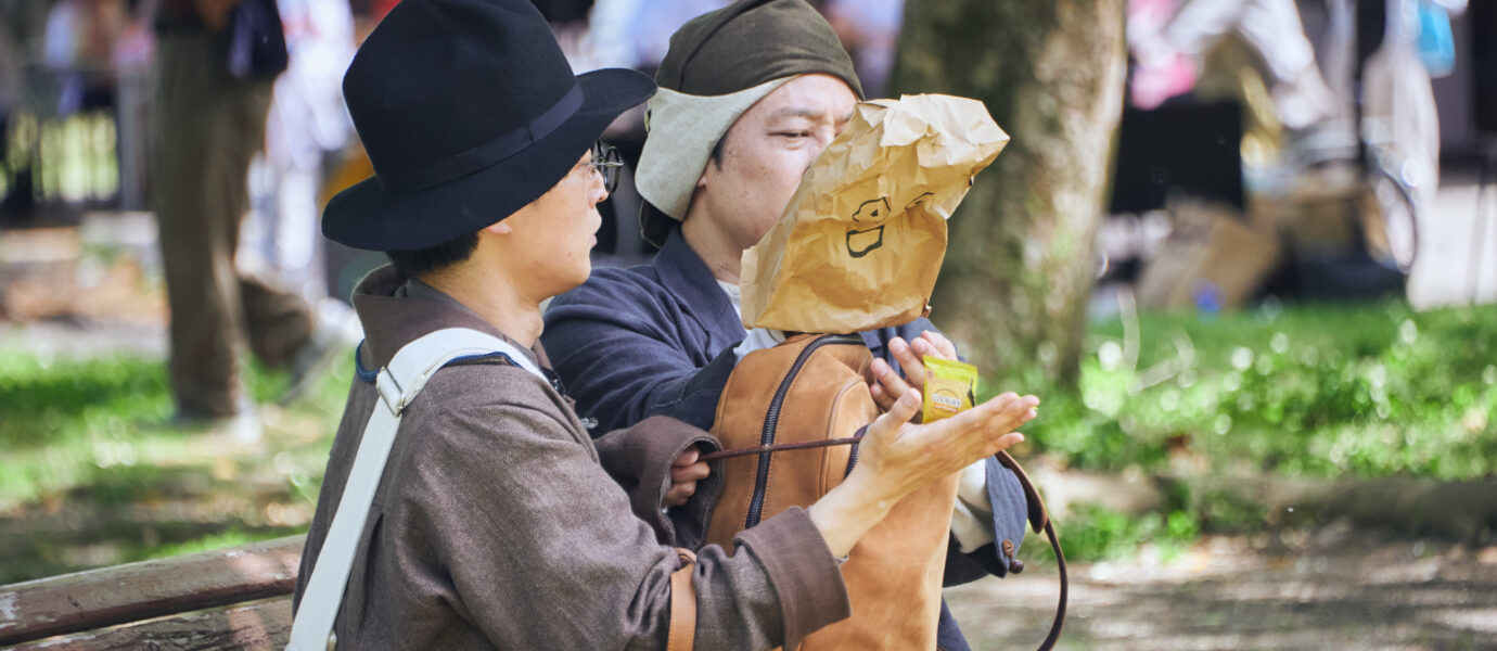 Two people sit on a park bench, one wearing a hat and the other wearing a paper bag over their face.
