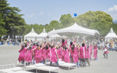 A group of people wearing pink tunics gather in a circle outdoors, reaching their arms towards a blue balloon floating above a white marquee.