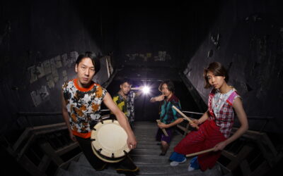 Four musicians holding taiko drums and drumsticks pose on a dark concrete stairway.